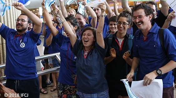 Large group of people dressed in blue, smiling, and waving Israeli flags.