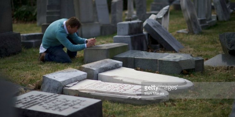 Man kneeling by several tombstones that have been destroyed in a cemetery.