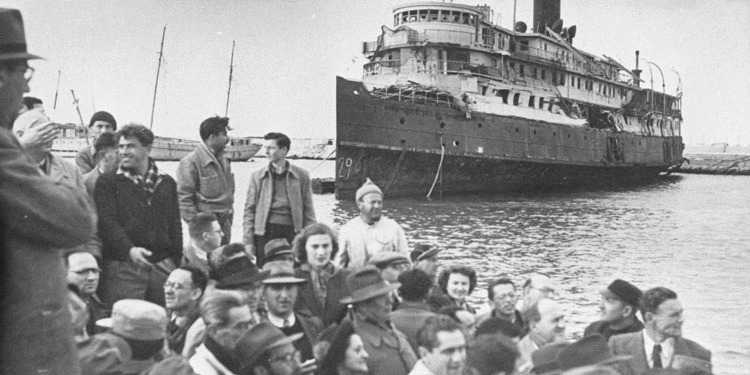 Black and white image of a ship coming into harbor with people waiting at the harbor.
