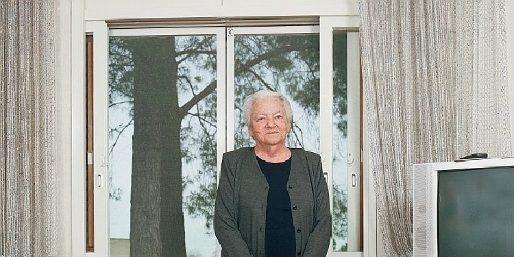 Elderly woman standing in front of large windows and glittery curtains staring straight ahead.