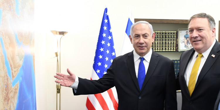 Bibi standing next to a map and two flags alongside another man in a suit.