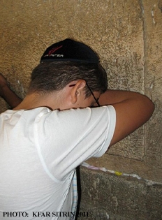 Young Jewish man wearing kippah leans on the Western Wall and bows his head in prayer. 