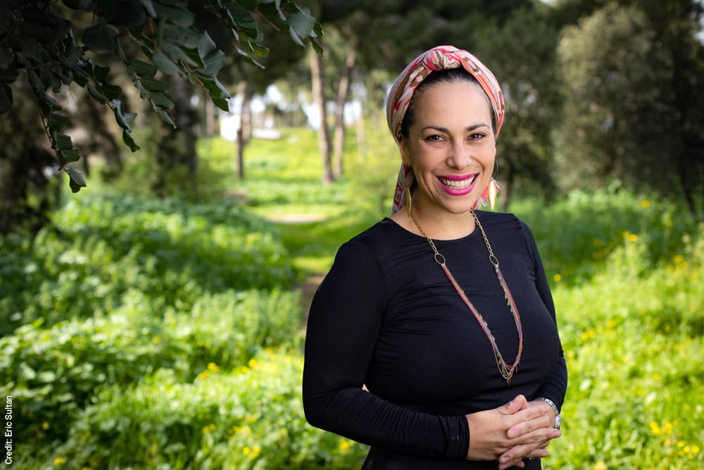 Yael Eckstein, wearing a black shirt, stands in a grassy wooded area smiling.