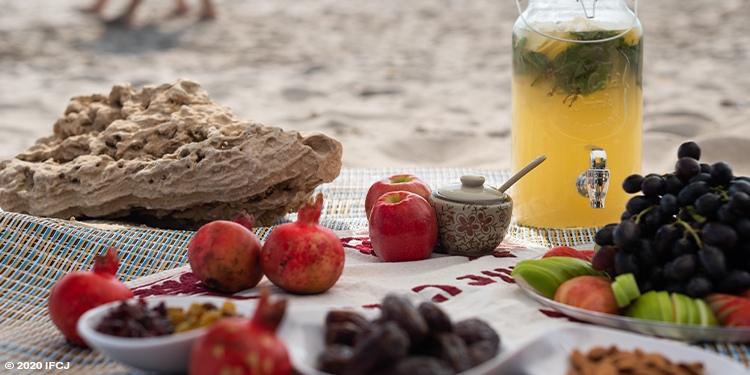 A picnic spread of a pitcher of juice, beehive, pomegranates, apples, and more fruit on a beach.