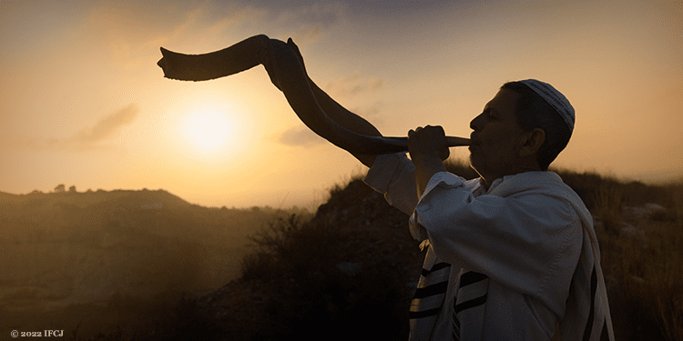 Man in a white robe blowing into a shofar against a sunset background.