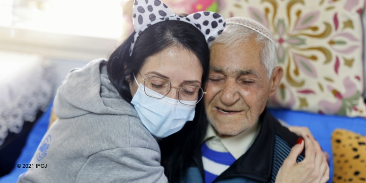 Woman in IFCJ sweatshirt hugging an elderly Jewish woman while they're sitting next to each other.