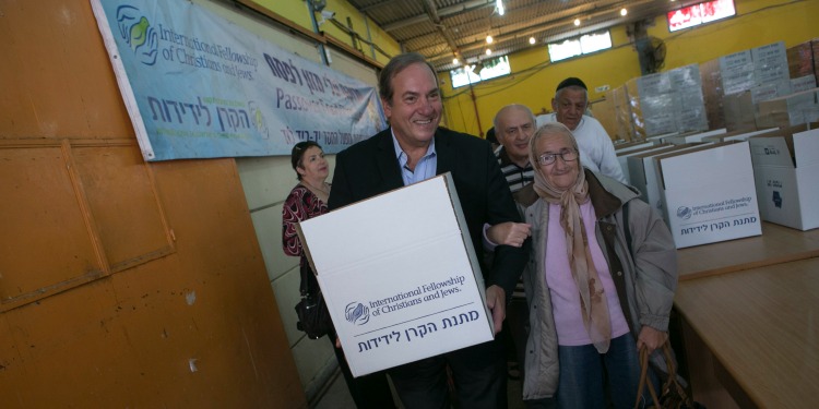Rabbi Eckstein holding an IFCJ branded food box as he's helping an elderly woman down a hallway.