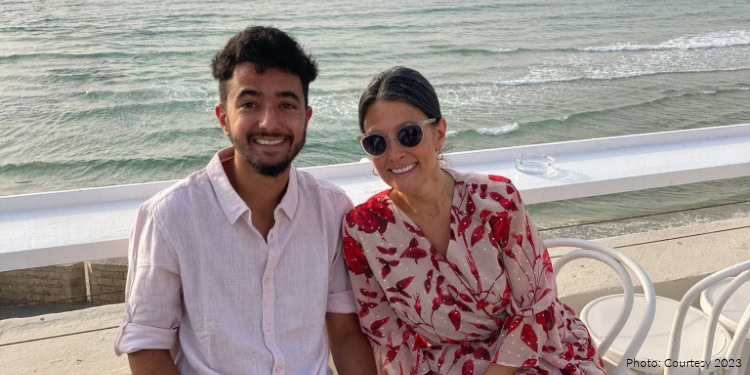 Happy couple sitting on a white bench by the ocean, smiling at the camera, with waves in the background, wearing casual summer clothes and sunglasses.