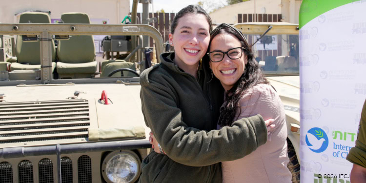 Two women embracing in front of a military vehicle, symbolizing hope and unity during difficult moments, aligned with the theme "This is Our Red Sea Moment.".