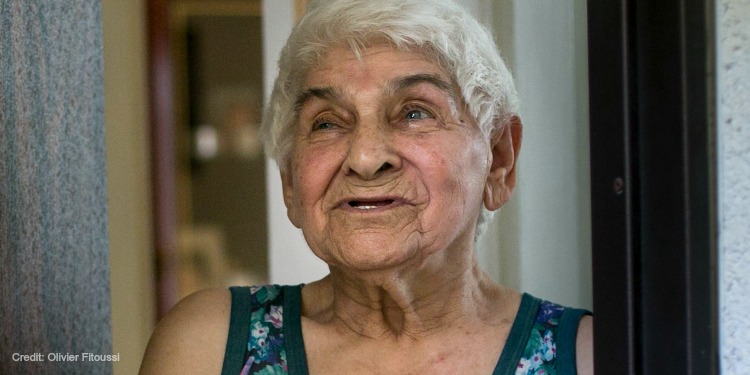 An elderly Jewish woman with white hair, blue eyes, and a blue floral top looking upwards.