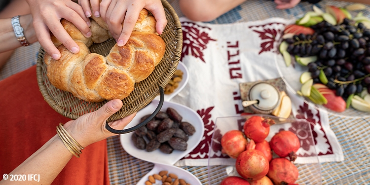 Hands gathered over a piece of bread with fruits in the background.