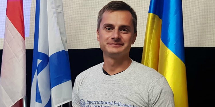 Oleksandr Milshtein an IFCJ recipient standing in front of 3 flags.