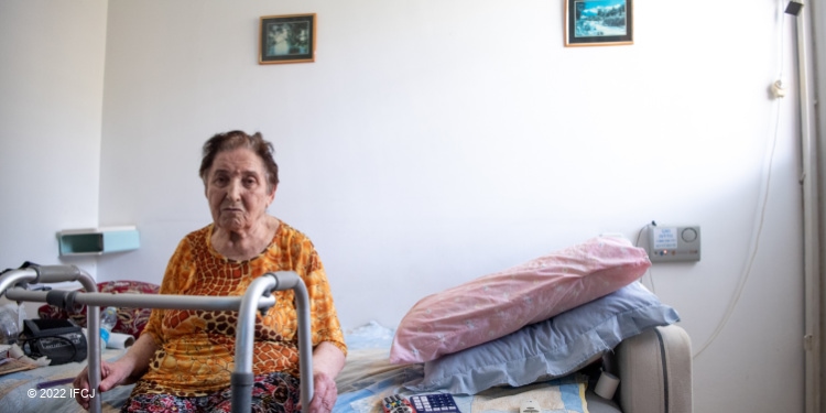 Elderly Jewish woman sitting on her bed with her walker in front of her.