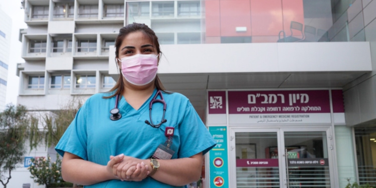Woman in scrubs and a pink mask standing in front of a hospital.