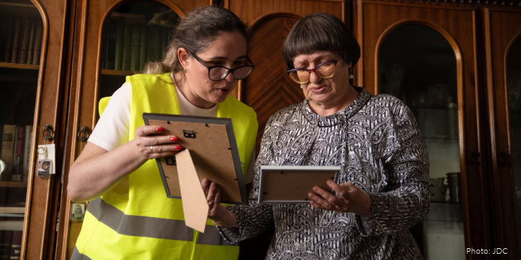 Two women, one in a yellow safety vest and the other in a patterned blouse, reviewing documents in an indoor setting with wooden furniture.