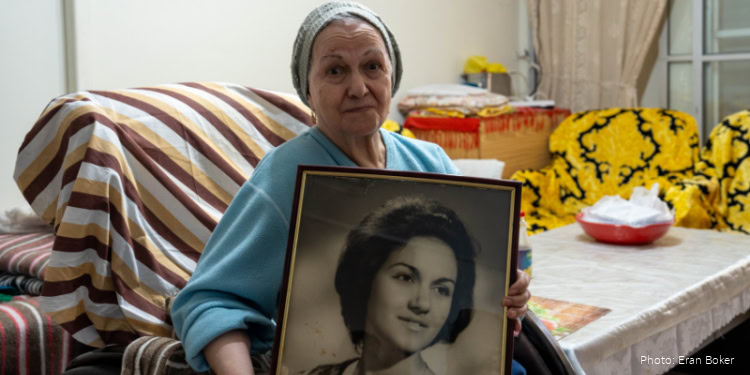 Elderly woman Holocaust survivor holding a framed black-and-white photo, sitting in a cozy living room with colorful cushions and a table, reflecting on her past.