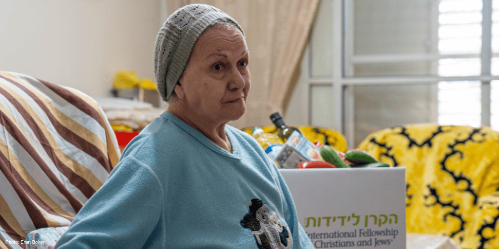 Elderly woman, sitting on couch, brown and white striped covering, blue sweatshirt, gray knitted hat, head covering, branded logo food box in back, IFCJ Korea - couch in rear with yellow covering - looking right