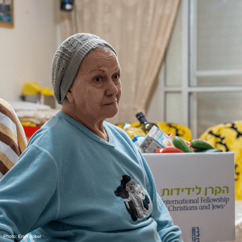 Elderly woman, sitting on couch, brown and white striped covering, blue sweatshirt, gray knitted hat, head covering, branded logo food box in back, IFCJ Korea - couch in rear with yellow covering - looking right