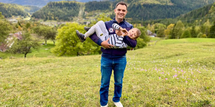 Man holding a smiling child in a green park with trees and mountains in the background.