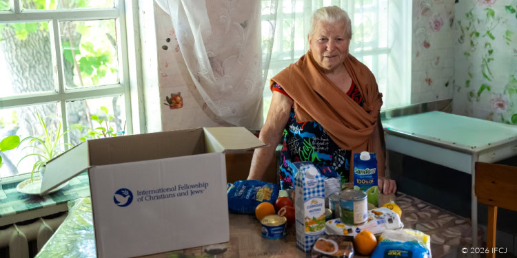 Elderly woman with white hair and a warm smile, standing at a table with food and supplies, in a cozy, well-lit room with floral wallpaper and large windows.