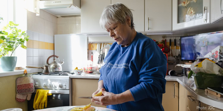 Elderly woman with gray hair wearing a blue sweater preparing food in a bright kitchen with natural light and modern appliances.