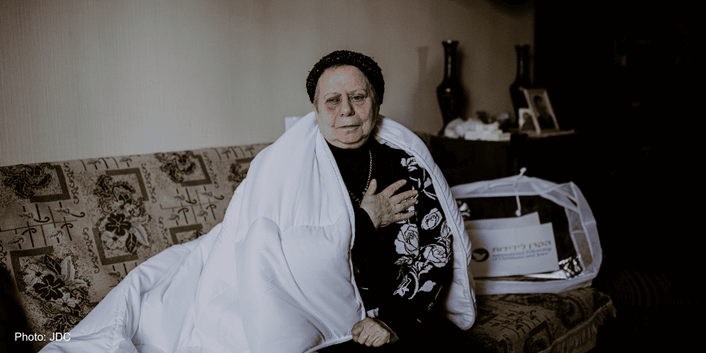 An elderly woman sitting on a sofa with a white blanket over her shoulders, holding her hand to her chest, expressing emotion or concern.