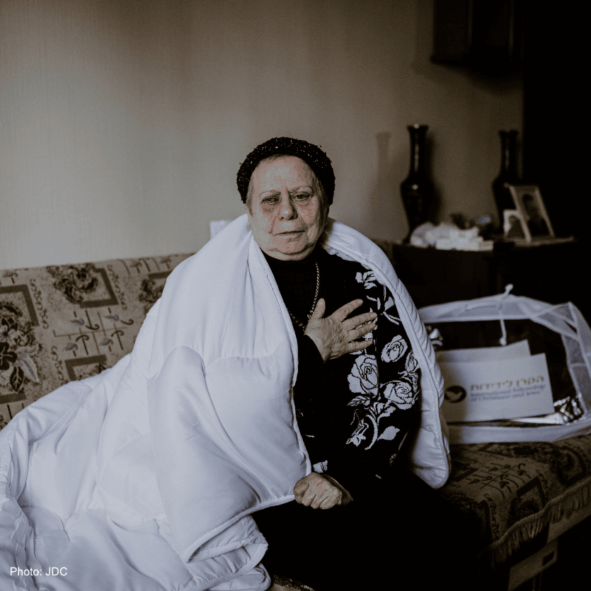 An elderly woman sitting on a sofa with a white blanket over her shoulders, holding her hand to her chest, expressing emotion or concern.
