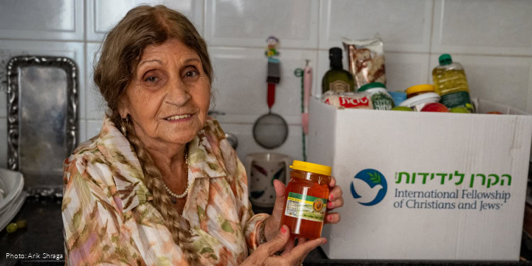 Visit to Esther David - With Dignity and Fellowship - Bat Yam, Israel - September 15, 2025 elderly woman holding jar of honey, cream floral dress, looking at camera, branded logo food box on kitchen counter