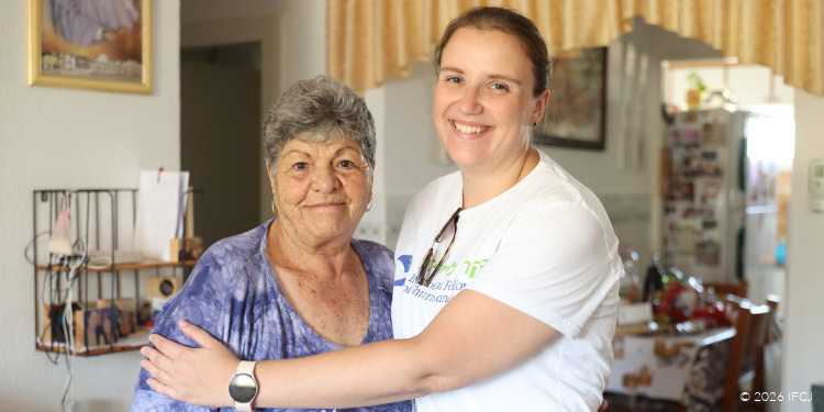 A young woman volunteer smiling and hugging an elderly woman in a cozy home setting, representing compassion and support from IFCJ.
