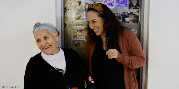 Visit to Hana Dahan - With Dignity and Fellowship - Tiberias, Israel - November 20, 2024 Yael Eckstein, black dress, brown sweater, talking with Hana Dahan, elderly woman, black sweater with red and white flowers, white shirt, gray headscarf - smiling, laughing - photos on metal door