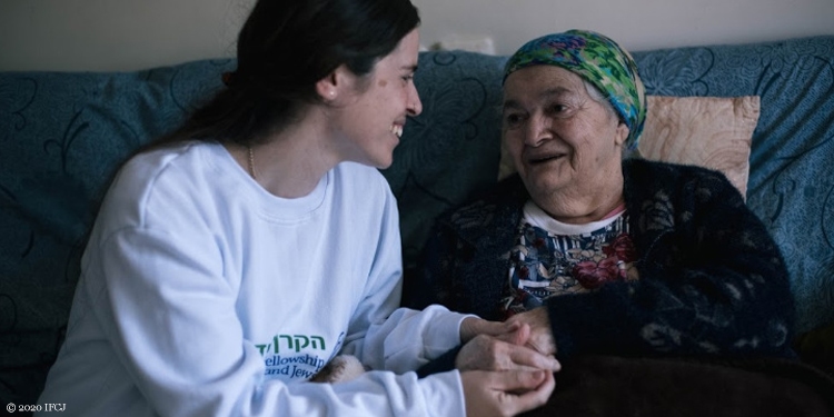 IFCJ staff member in an IFCJ crew neck holding the hand and smiling at an elderly Jewish woman.
