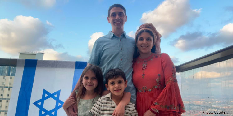 Iair and Sara Fogel and their children, Clara and Jaim, olim family from Argentina. family standing on rooftop overlooking Israeli city, Israel flag draped on railway, smiling, happy