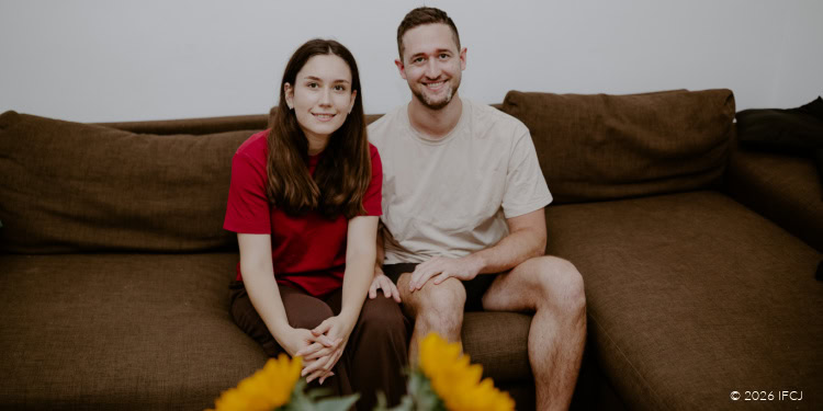 Ilia Fedorov and Ekaterina Litvinova - Russia Aliyah - Tbilisi, Georgia - October 20, 2025 young adult couple sitting on brown couch looking at camera - young woman, red t-shirt - young man, beard, tan t-shirt - sunflowers
