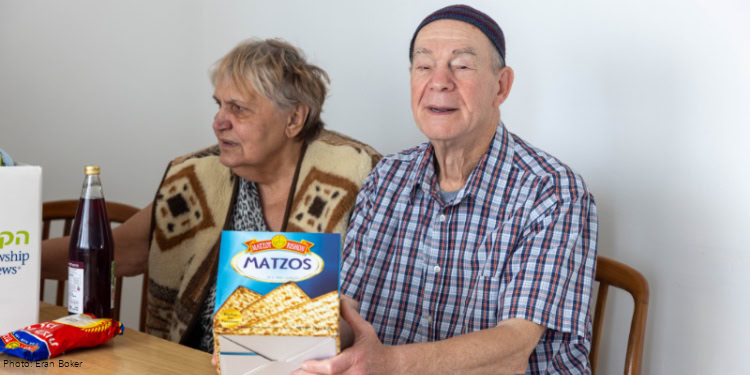 Visit to Illia and Raisa Farberov - With Dignity and Fellowship – Passover 2025 - Haifa, Israel - March 25, 2025 elderly man in red, black, white plaid shirt, black cap sitting at table -box of matzah - elderly woman, animal print dress, tan and brown vest over her shoulders - food items on table.