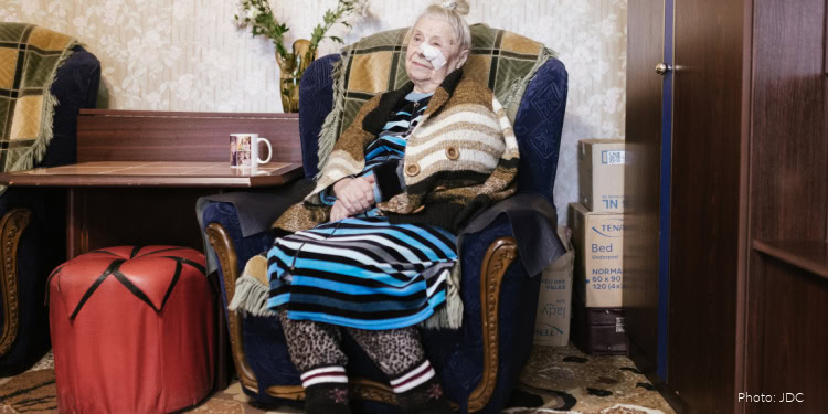 Inna Kolyadova - JDC - Kishinev, Moldova - November 13, 2024 elderly woman, blue and black striped sweater, brown and beige striped sweater, white bandage, navy blue chair,green and yellow blanket