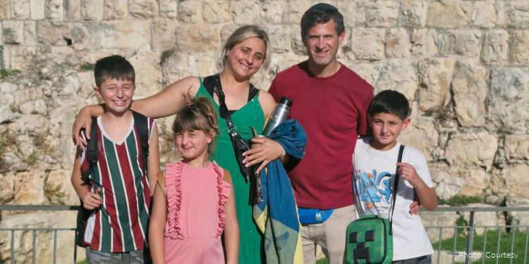 A happy family of five, including two children and two adults, smiling and embracing each other outdoors in front of a stone wall, symbolizing unity and support.