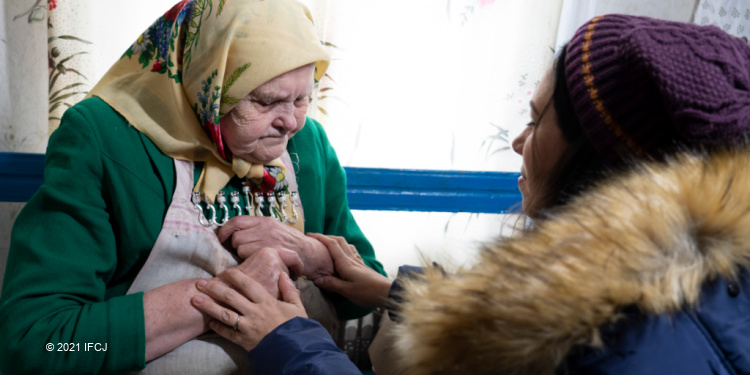 Katerina Palamerchuk, age 94, Yael Eckstein kneels in front of and holds hands of elderly woman wearing multiple layers of clothing and yellow scarf on her head,