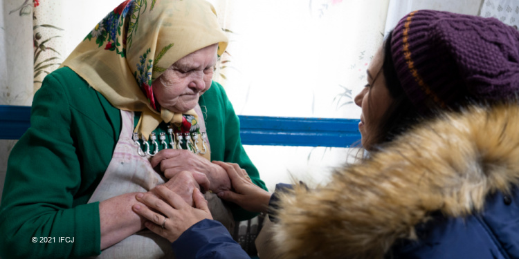 Yael Eckstein kneels in front of and holds hands of elderly woman wearing multiple layers of clothing and yellow scarf on her head,