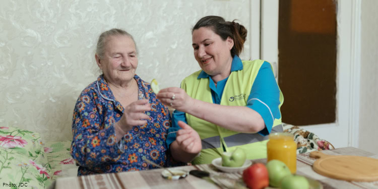 Visit to Khona Belaya - JDC - Bender, Moldova - July 10, 2025 woman in turquoise blue shirt, yellow safety vest holding up slice of apple for elderly woman in blue pajamas with floral pattern, sitting in a bed - green sheets with pink flowers - table with brown and beige tablecloth, plate of apples, jar of honey