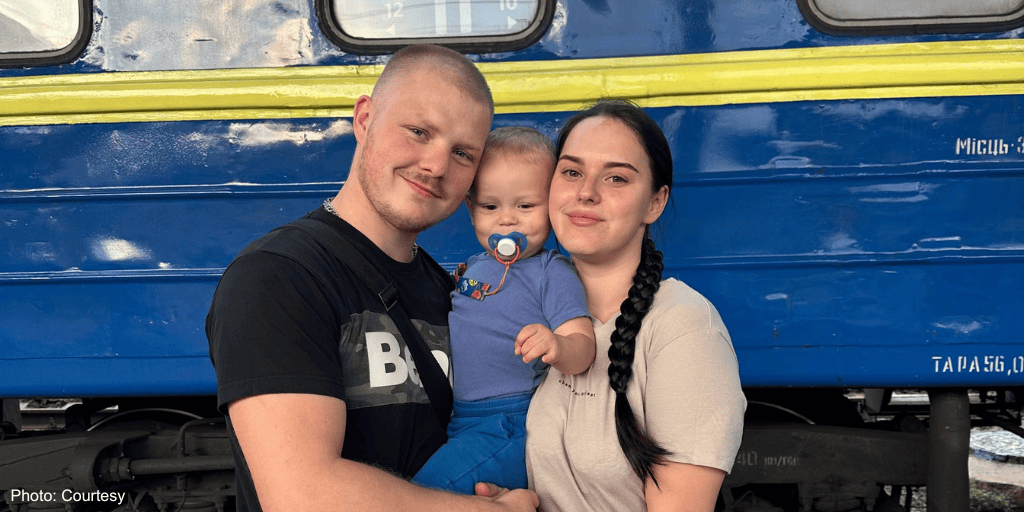 A happy family of three, including a young child with a pacifier, smiling and embracing each other in front of a blue train, symbolizing hope and kindness.