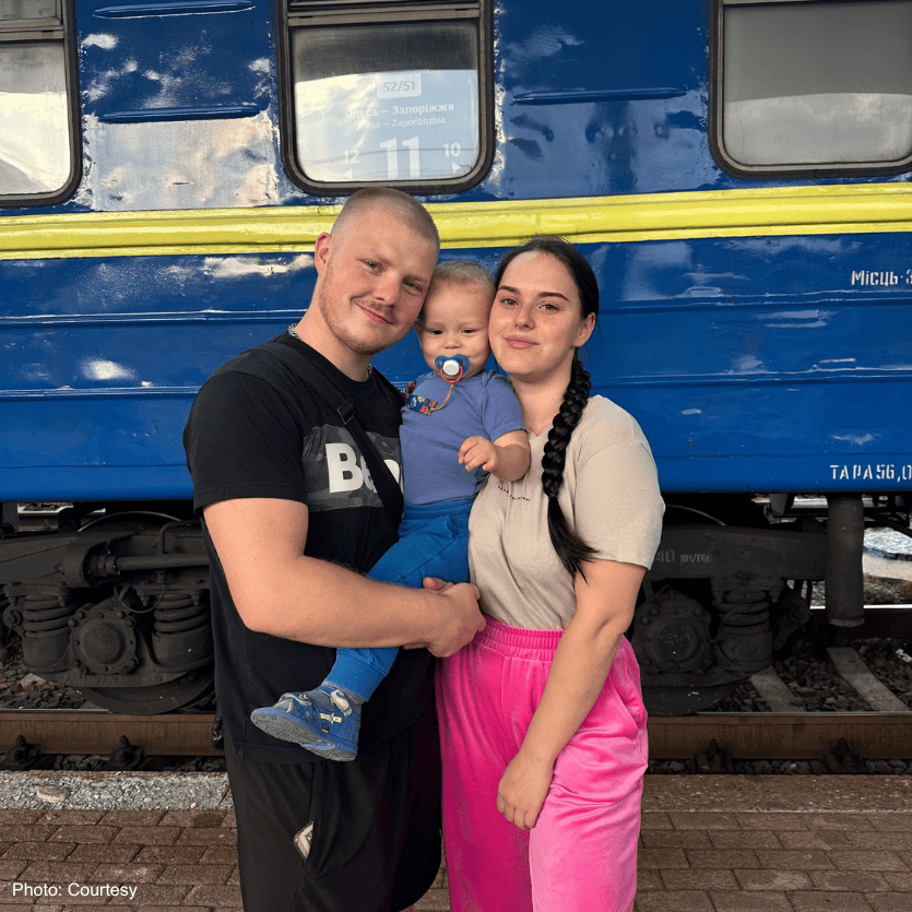 A happy family of three, including a young child with a pacifier, smiling and embracing each other in front of a blue train, symbolizing hope and kindness.