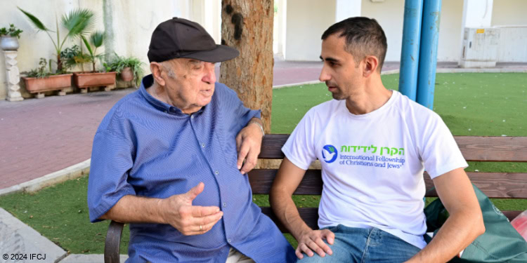 Yad B’Yad, elderly man sit with Gadi Dan (IFCJ Staff) outside in courtyard, branded logo tshirt