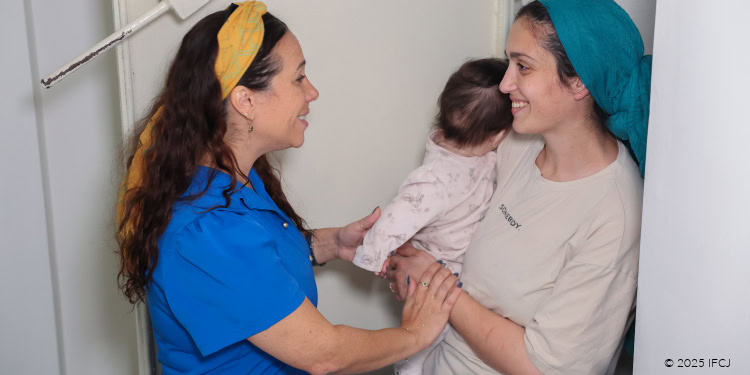 Visit with Lian Borochov and Baby Avigayil - Survivors of Iranian Rocket Attack (June 2025) - November 18, 2025 Yael Eckstein, blue dress, yellow headband - standing behind heavy white door to safe room - young woman, turquoise head scarf, cream t-shirt - holding baby Avigayil in pink outfit - smiling at Yael - Yael touching her arm, holding baby's hand, caring, concern