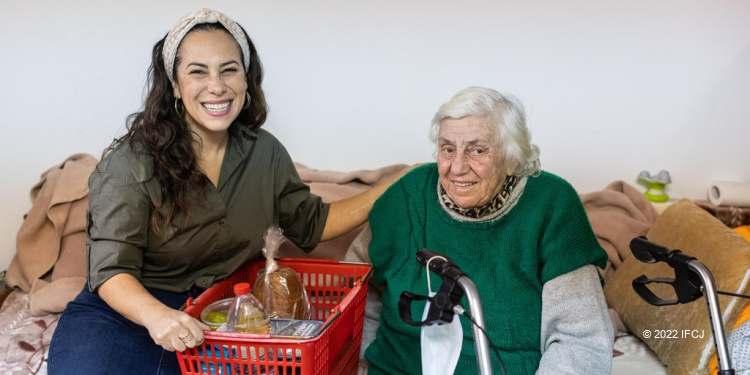 Yael Eckstein sitting with an elderly Jewish woman and a basket of donated food on a couch.