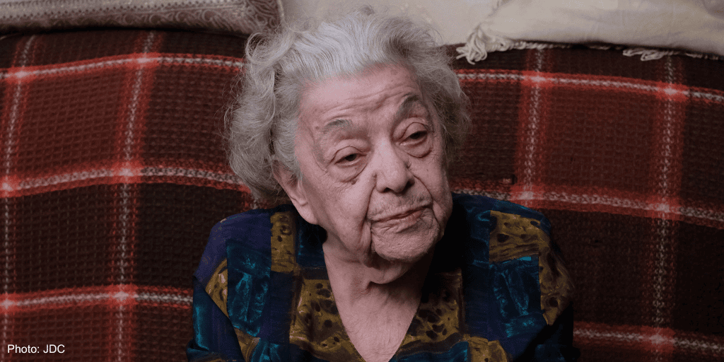 Elderly woman with gray hair and a thoughtful expression sitting on a red plaid couch, wearing a colorful blouse, in a cozy home setting.