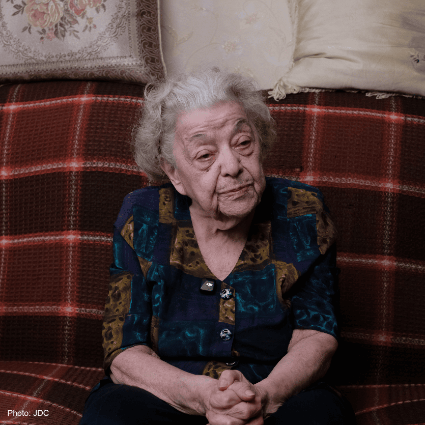 Elderly woman with gray hair and a thoughtful expression sitting on a red plaid couch, wearing a colorful blouse, in a cozy home setting.