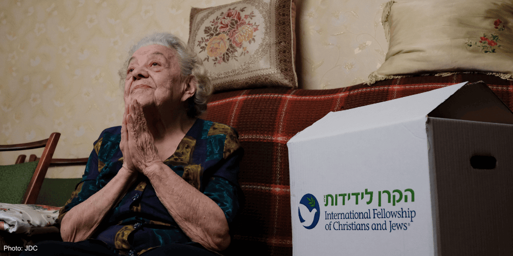 elderly woman, blue and brown blouse - sitting on couch, red and brown plaid blanket - branded logo food box - looking up, hands clasped in prayer
