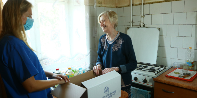 Elderly woman smiling at an IFCJ staff member in a blue shirt and mask while receiving an IFCJ food box.