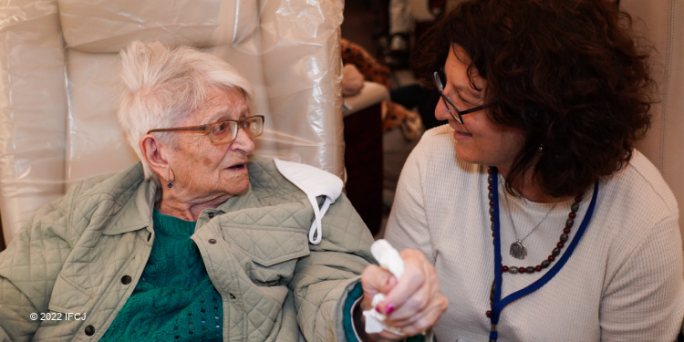 Elderly Jewish woman in green holding the hand of a female IFCJ staff member.