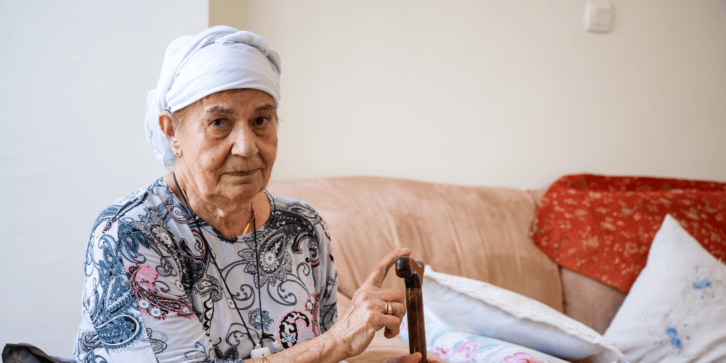 Marcel Bar David visit to elderly woman's home. elderly woman, blue and green paisley dress, headscarf, sitting in black armchair, holding cane. Brown couch, orange blanket, white pillows